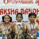 Three smiling schoolchildren in beige uniforms hold festive craftsa red rakhi toward the camera with a banner reading observation of raksha bandhan in the background behind them